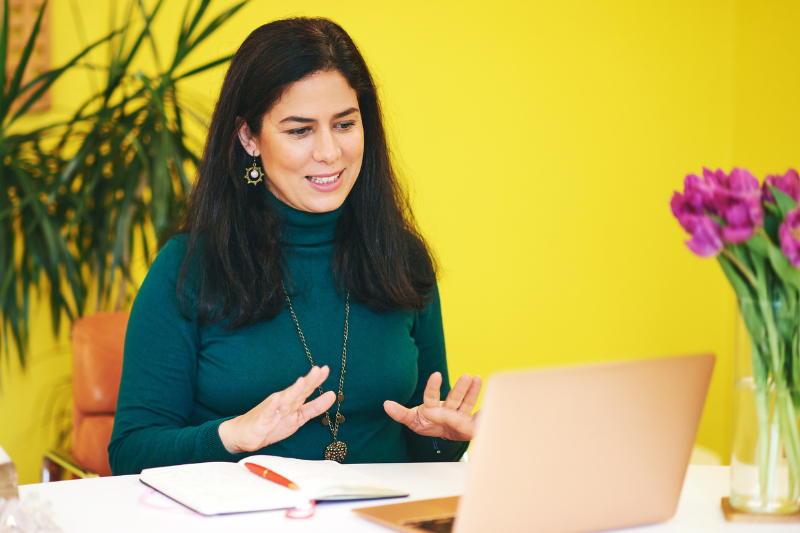 Woman speaking to someone on video call on laptop in front of yellow wall
